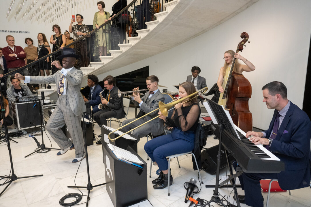 Dandy Wellington, a Black man in a gray striped suit dances in front of his seven piece jazz band in the Museum's rotunda. Behind them, guests in 1920s dress enjoy the performance. 